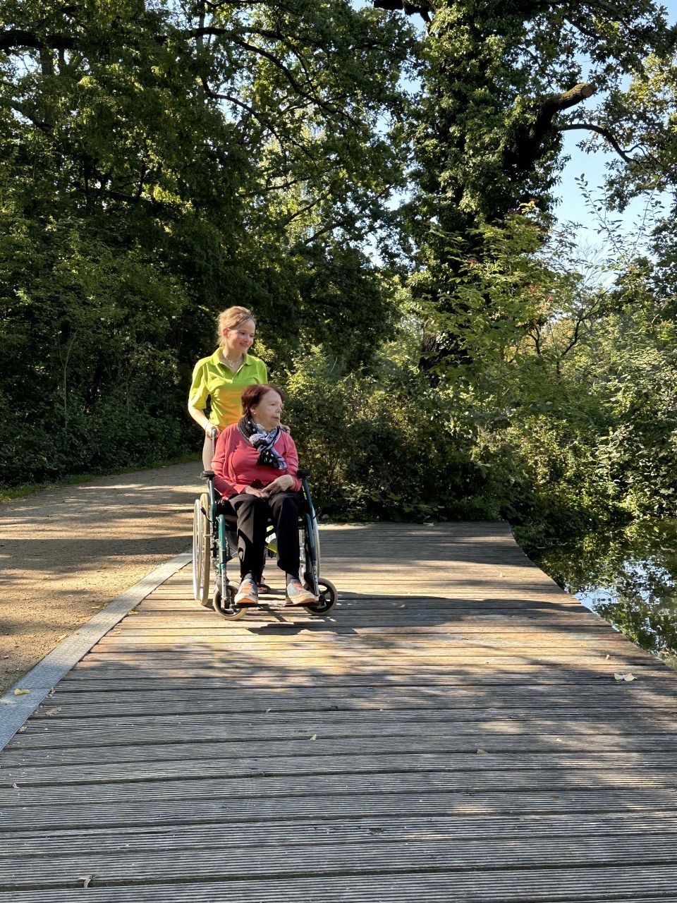 Eine Person im Rollstuhl wird von einer anderen Person auf eine Holzbrücke geschoben, in der Nähe von Bäumen und Wasser.