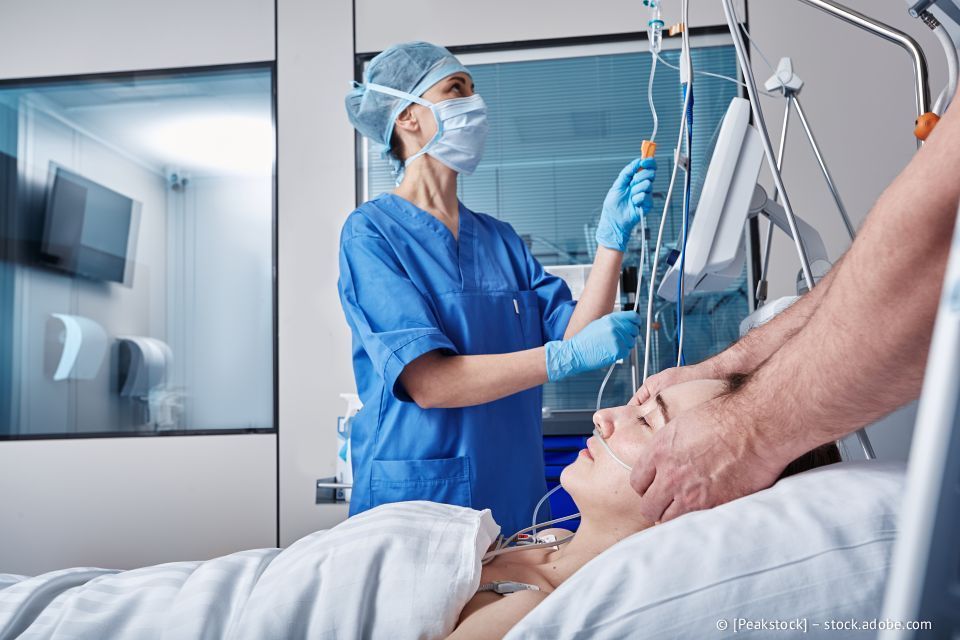 Eine Krankenschwester justiert den intravenösen Tropf eines Patienten im Krankenhausbett; die Hände einer anderen Person stützen den Kopf des Patienten.