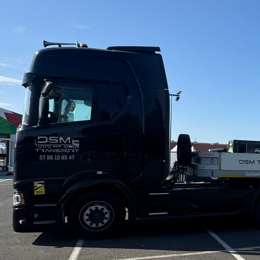 Un camion à plateau noir garé sur un parking, sous un ciel bleu azur.