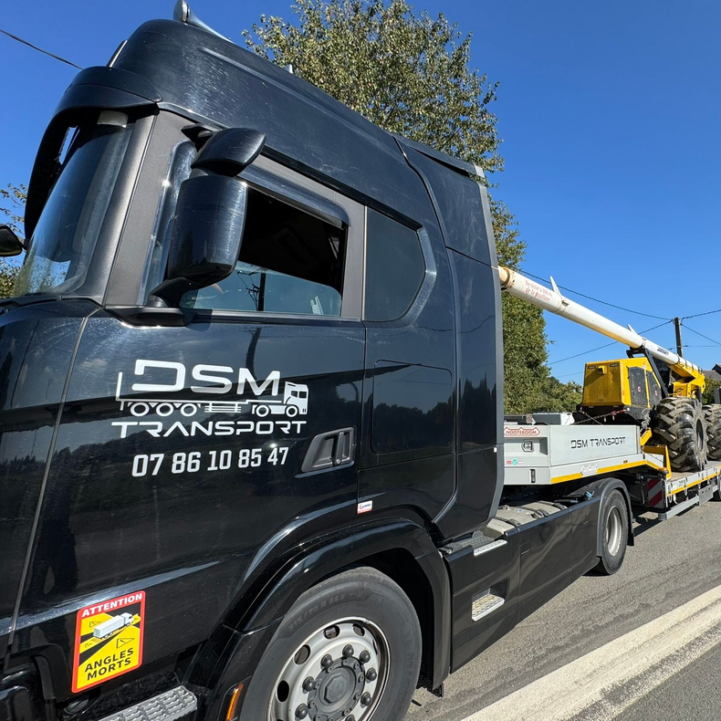 Une dépanneuse noire transporte un engin de chantier jaune sur une route sous un ciel bleu.