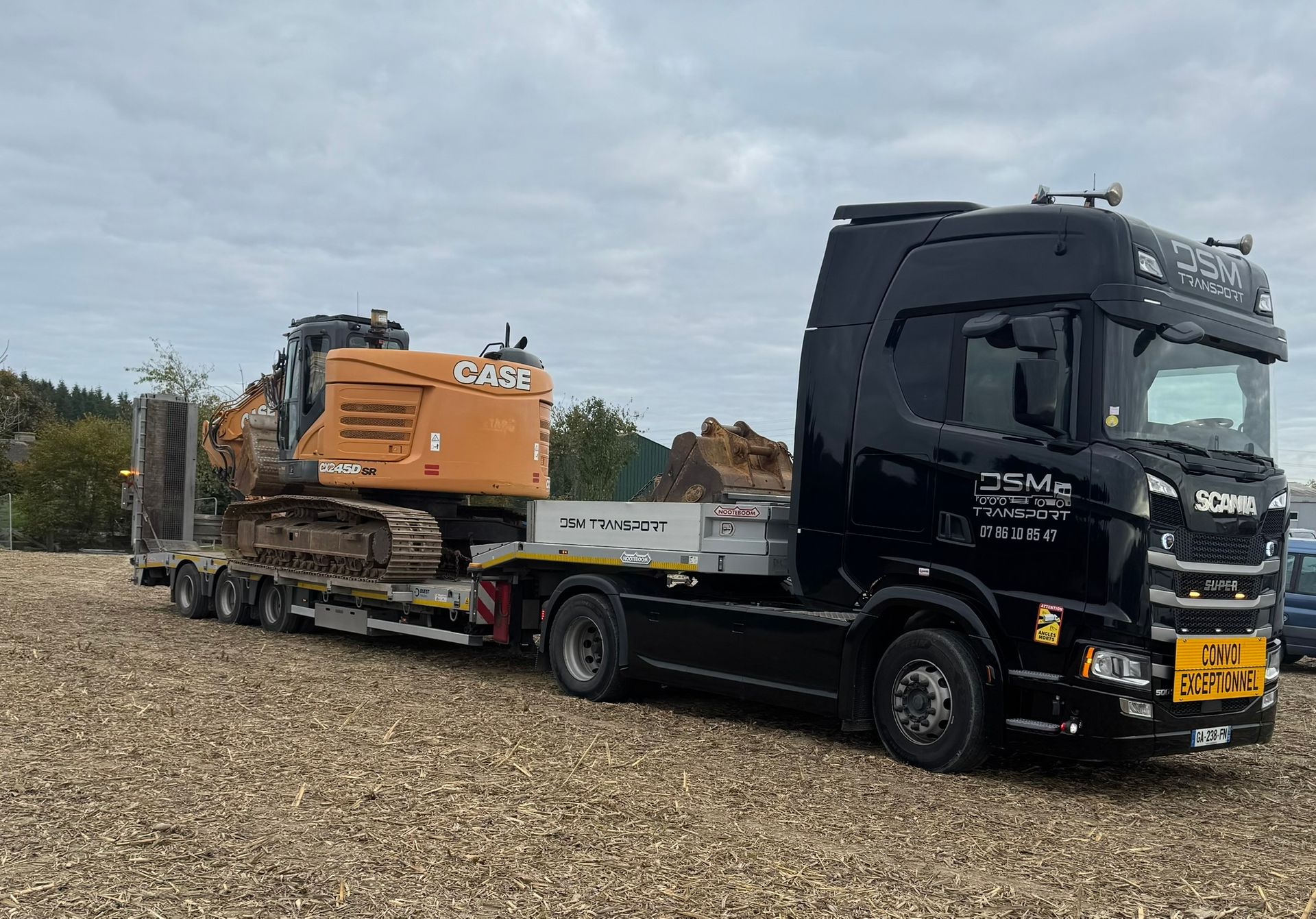 Un camion plateau noir transportant une excavatrice orange sur un terrain gravillonné.