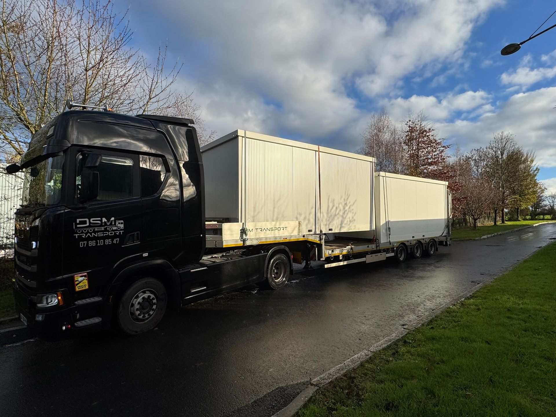 Un camion noir tirant deux remorques blanches sur une route mouillée, sous un ciel nuageux.
