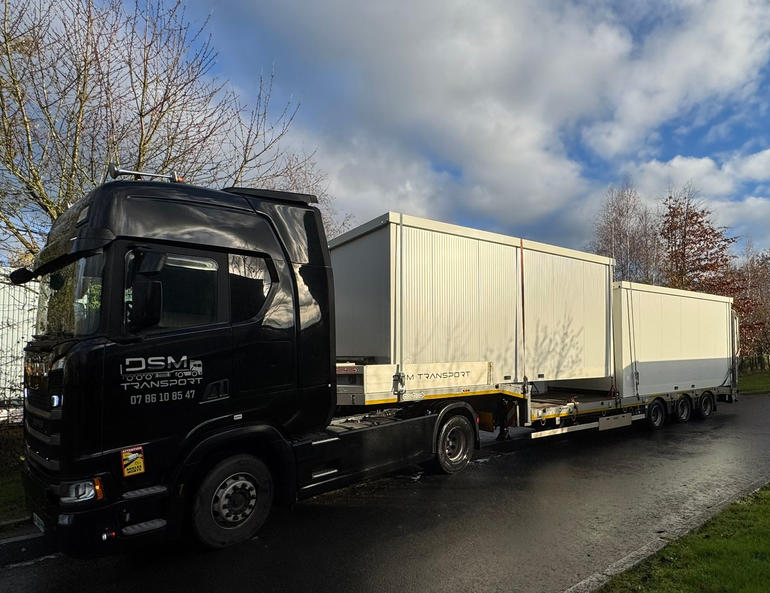 Camion de marchandises noir transportant deux conteneurs blancs sur une route mouillée, sous un ciel nuageux.
