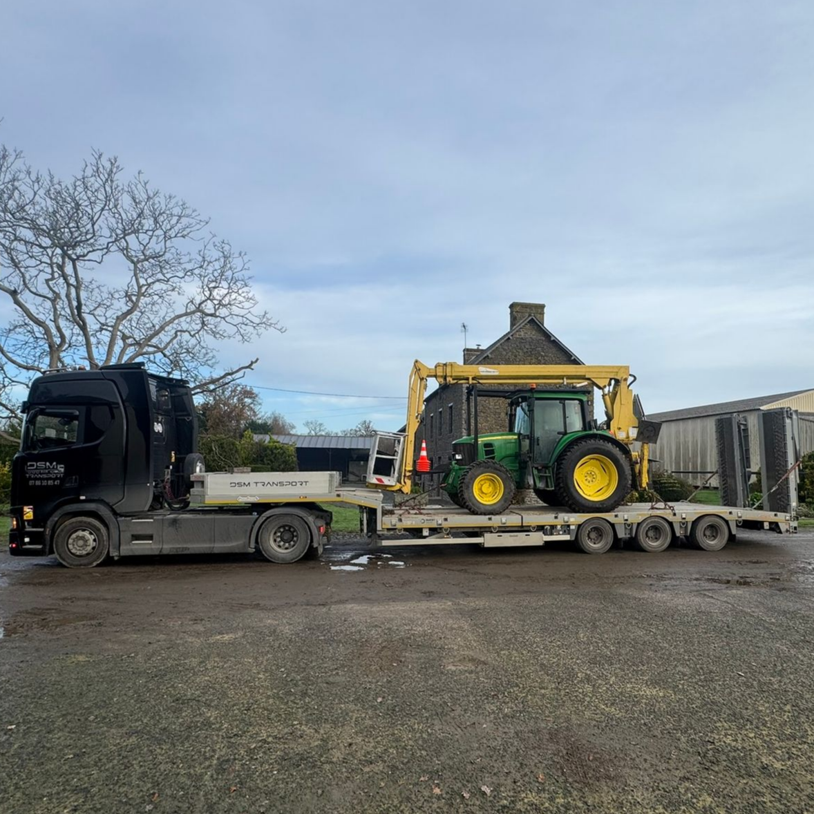 Camion plateau transportant un tracteur vert et jaune au bord d'une route de campagne.
