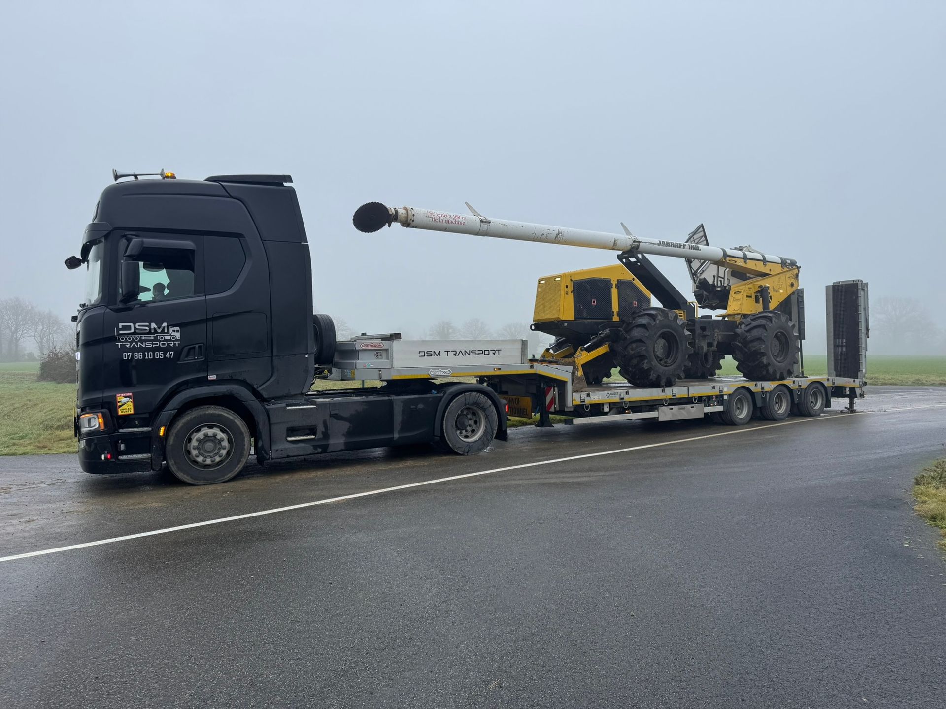 Un camion plateau noir transportant une machine agricole jaune sur une route mouillée.