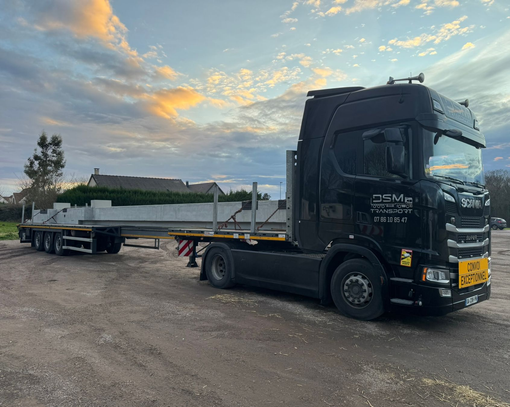 Un camion plateau noir tracte une longue poutre blanche dans une gravière, sous un ciel nuageux au coucher du soleil.