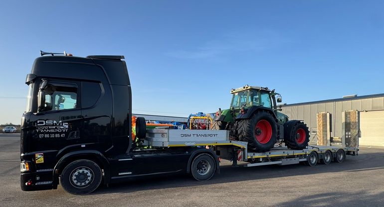 Un camion de dépannage noir à plateau transportant un tracteur vert sur un site industriel ensoleillé.