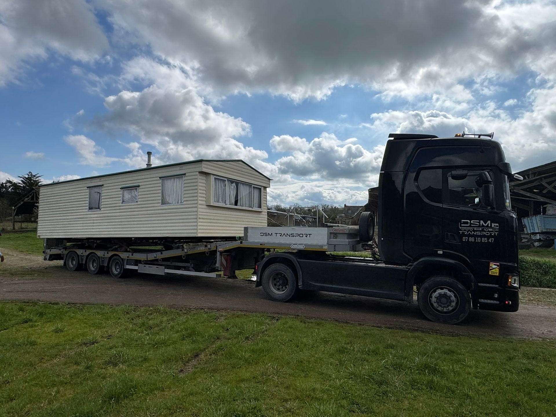 Un camion noir tracte un mobile-home blanc sur une remorque, sous un ciel nuageux.