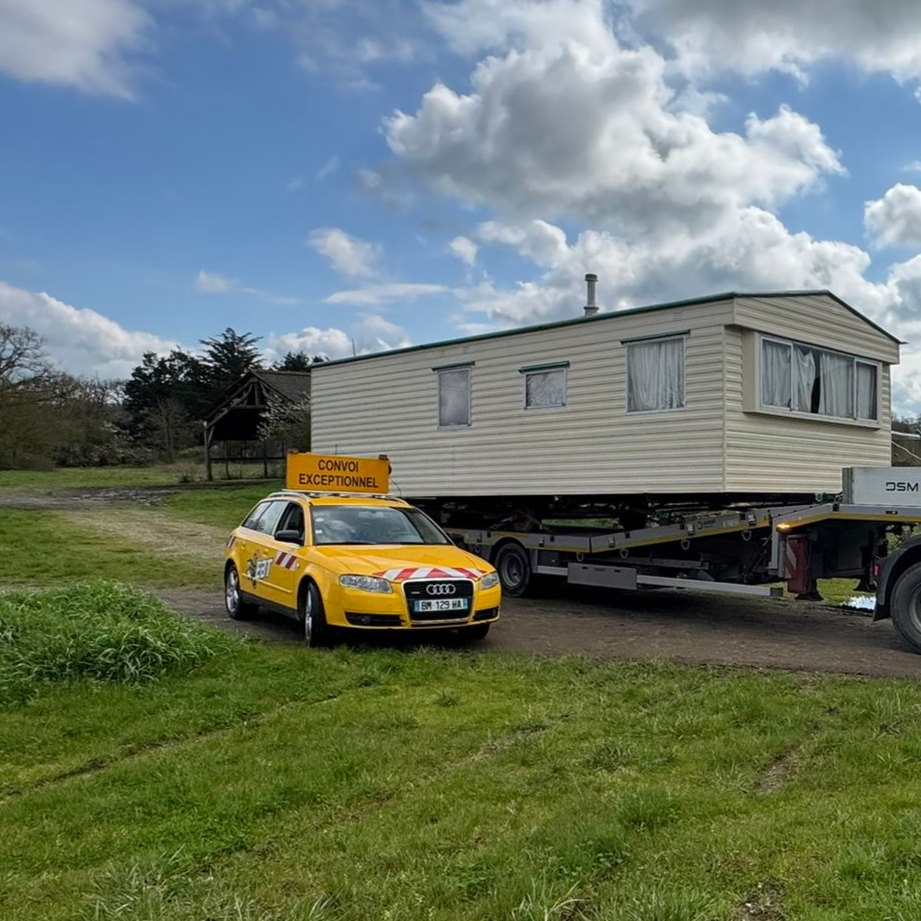 Une voiture jaune avec l'inscription Convoi Exceptionnel est garée devant un mobile home beige.