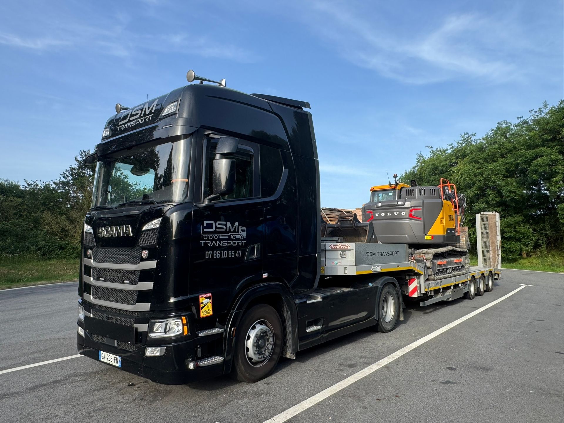 Un camion noir transporte un rouleau compresseur sur une remorque à plateau en bordure d'autoroute.