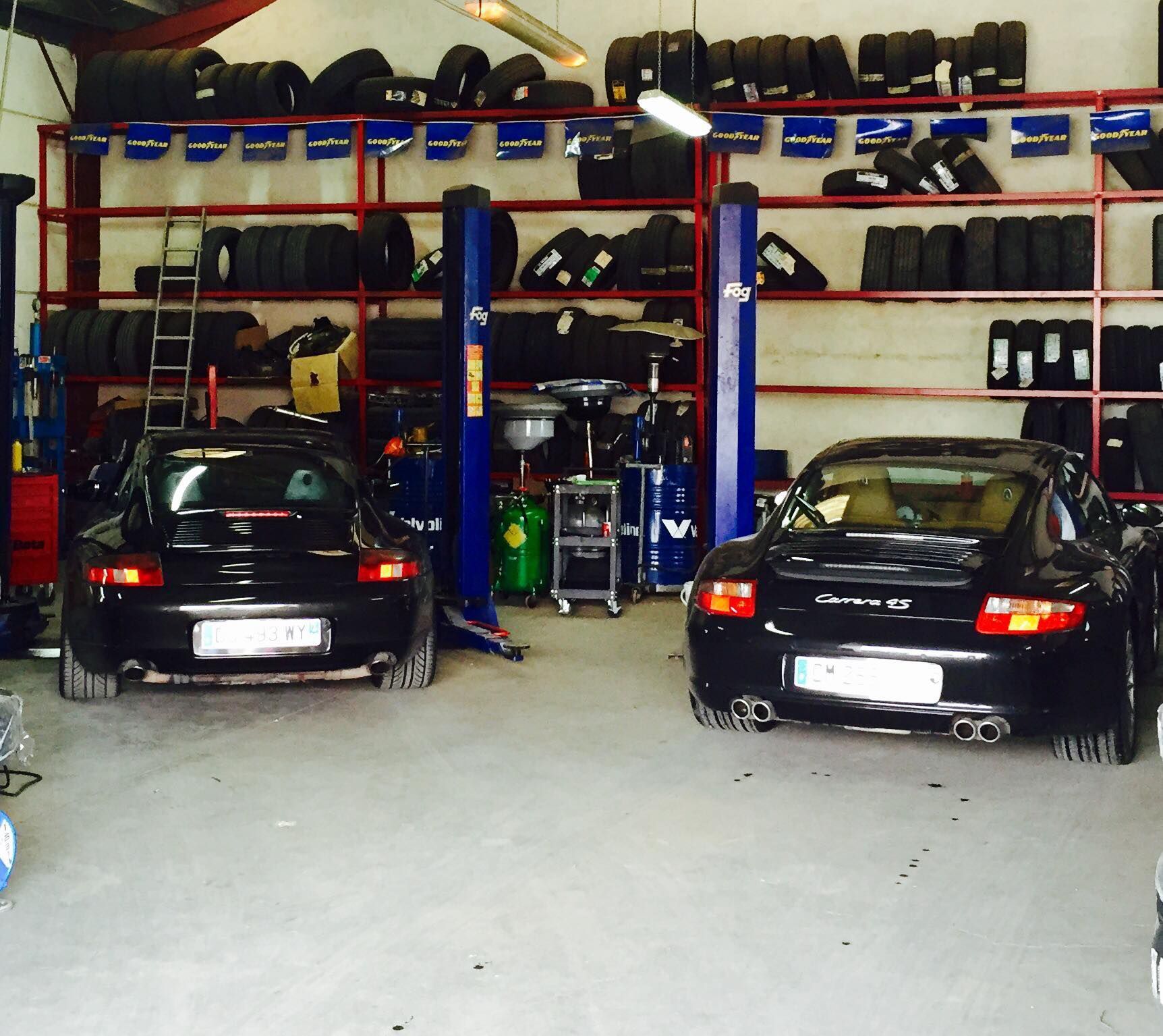 Deux voitures Porsche noires à l'intérieur d'un garage avec des pneus sur des étagères, des ascenseurs et des outils visibles.