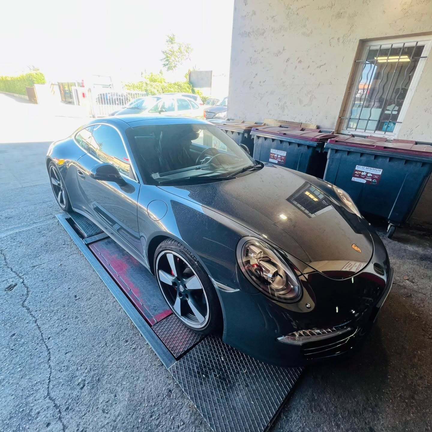 Voiture de sport Porsche gris foncé sur un ascenseur, devant des poubelles, à l'extérieur.