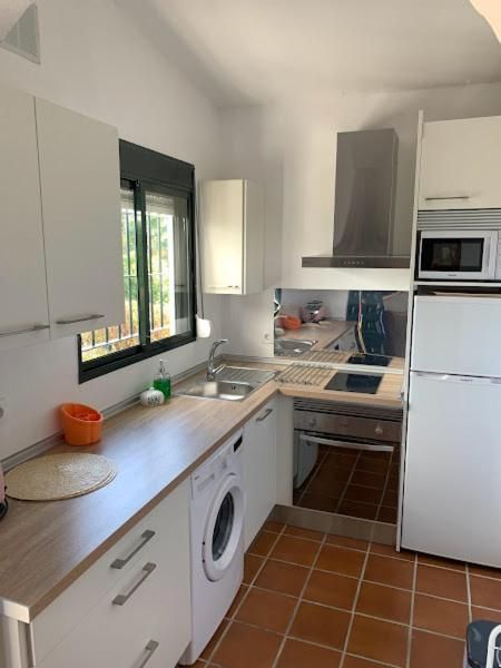 Cocina en forma de L con gabinetes blancos, encimeras claras, electrodomésticos y ventana. Suelo de baldosas marrones.