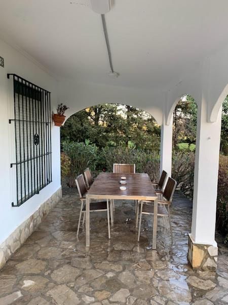 Un patio cubierto con una mesa de madera y seis sillas. A la izquierda, un muro con una ventana enrejada; árboles al fondo.