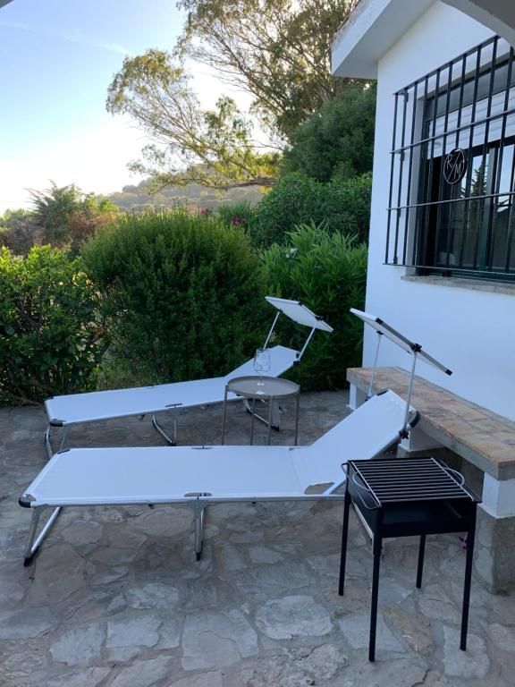 Dos sillones blancos, una mesa pequeña y una parrilla se encuentran en un patio de piedra al lado de un edificio.