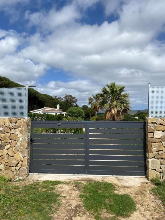 Puerta de metal gris situada en un muro de piedra, que conduce a una casa con palmeras bajo un cielo nublado.