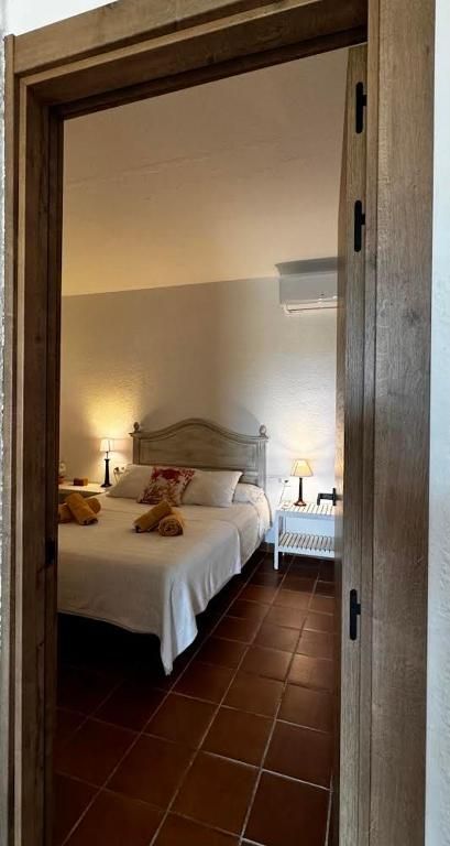 Interior del dormitorio visto a través de la puerta; cama blanca, piso de terracota, marco de puerta de madera, dos lámparas y aire acondicionado.