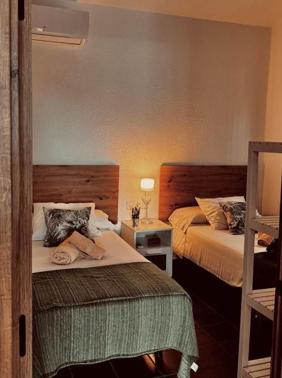 Dos camas individuales en una habitación con mesita de noche y lámpara; paredes blancas, cabeceros de madera.