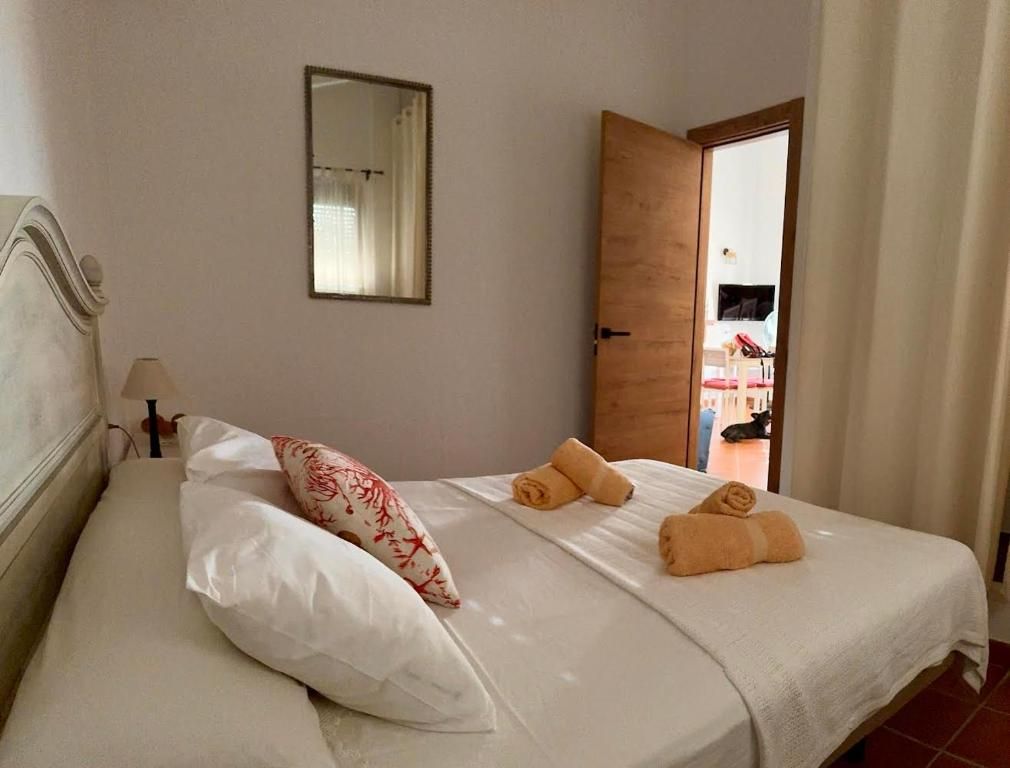 Dormitorio con cama blanca, toallas naranjas y una almohada decorativa. Puerta entreabierta que da a otra habitación con muebles.