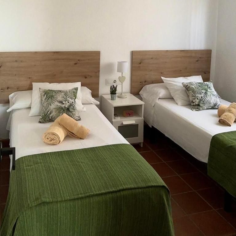 Dos camas individuales en una habitación con paredes blancas. Cada cama tiene cabecero de madera, sábanas blancas y una manta verde.