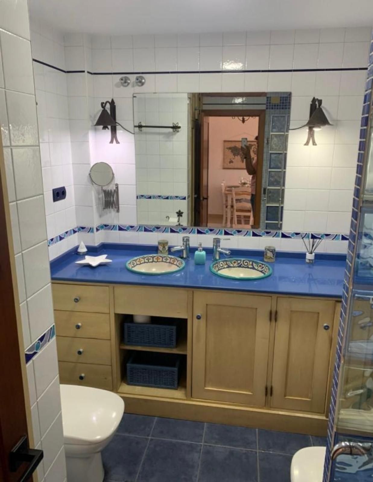Baño con encimeras azules, muebles de madera clara y dos lavabos, con espejo y piso de baldosas azules.