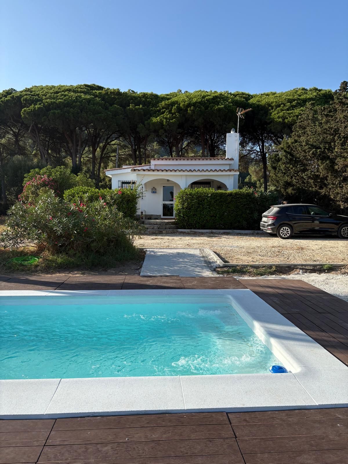 Piscina frente a una cabaña blanca con un coche aparcado cerca, rodeada de árboles y un cielo azul claro.