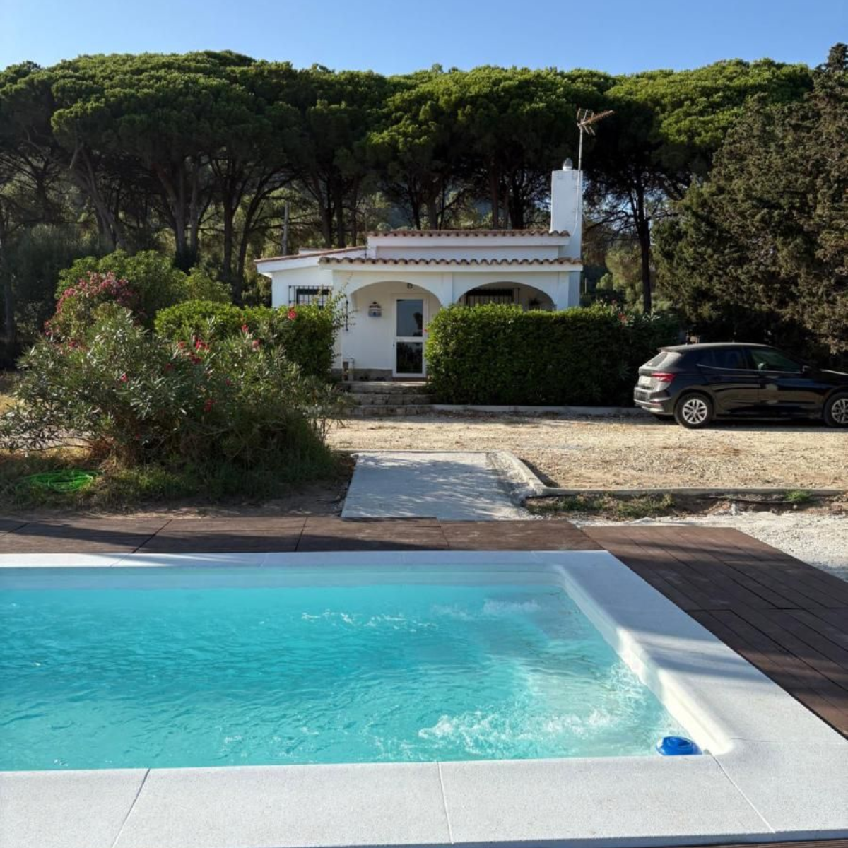 Piscina frente a una casa blanca con un coche aparcado cerca, rodeada de árboles.