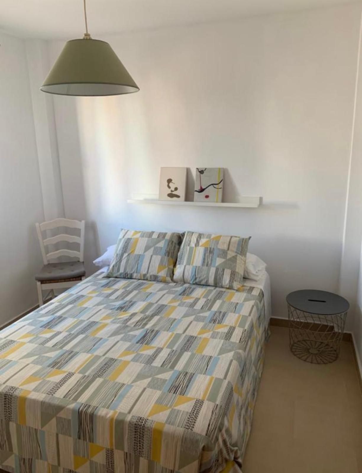 Dormitorio con cama, ropa de cama estampada, mesa pequeña, silla y arte en paredes blancas.