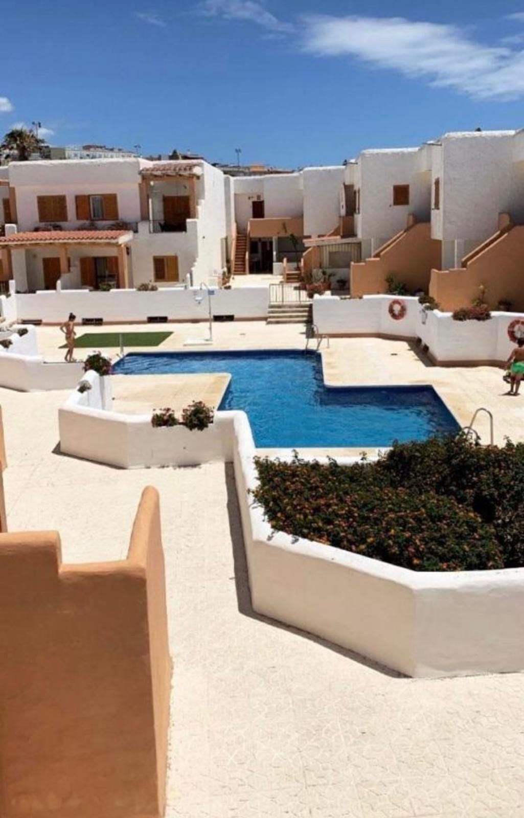 Piscina en complejo de estuco blanco con agua azul, rodeada de patios, plantas y edificios bajo un cielo soleado.