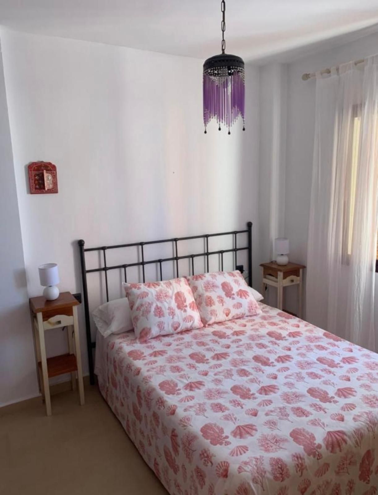 Dormitorio con estructura de cama de metal negro, dos mesitas de noche y ropa de cama con estampado rosa, bajo una lámpara colgante de color violeta.