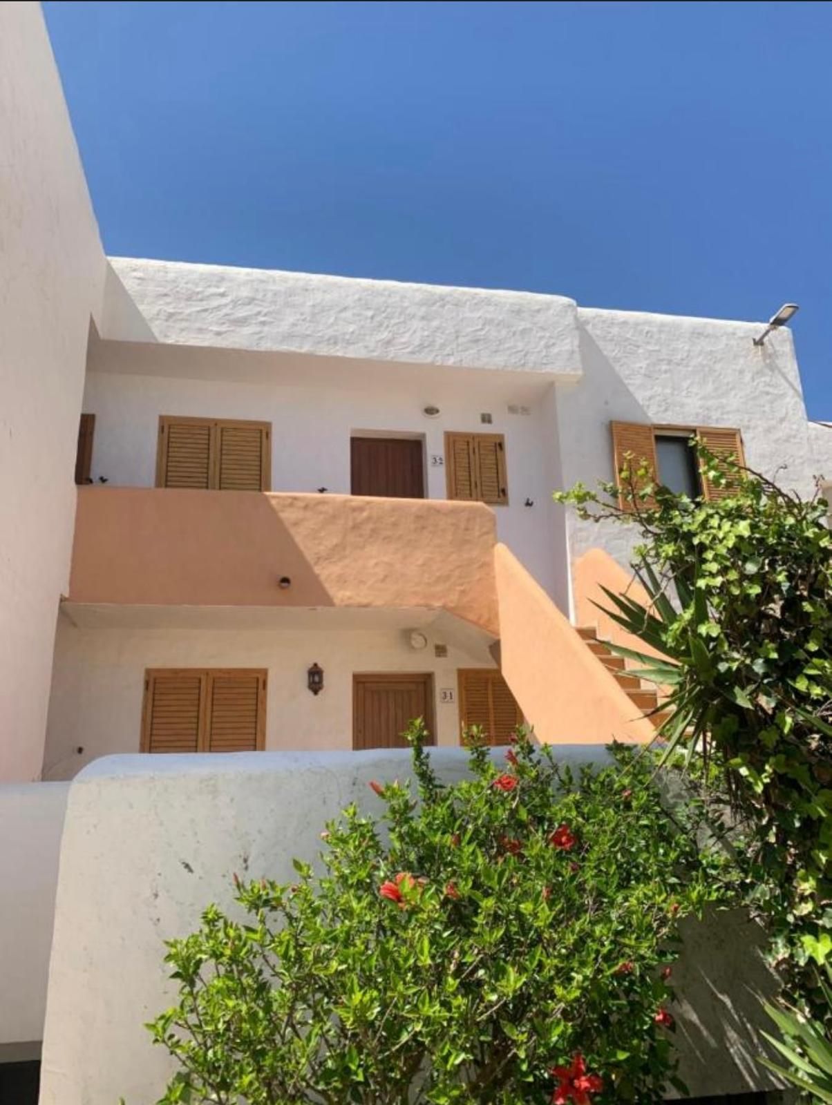Edificio de estuco blanco con detalles en color durazno, contraventanas de madera y exuberante vegetación bajo un cielo azul.