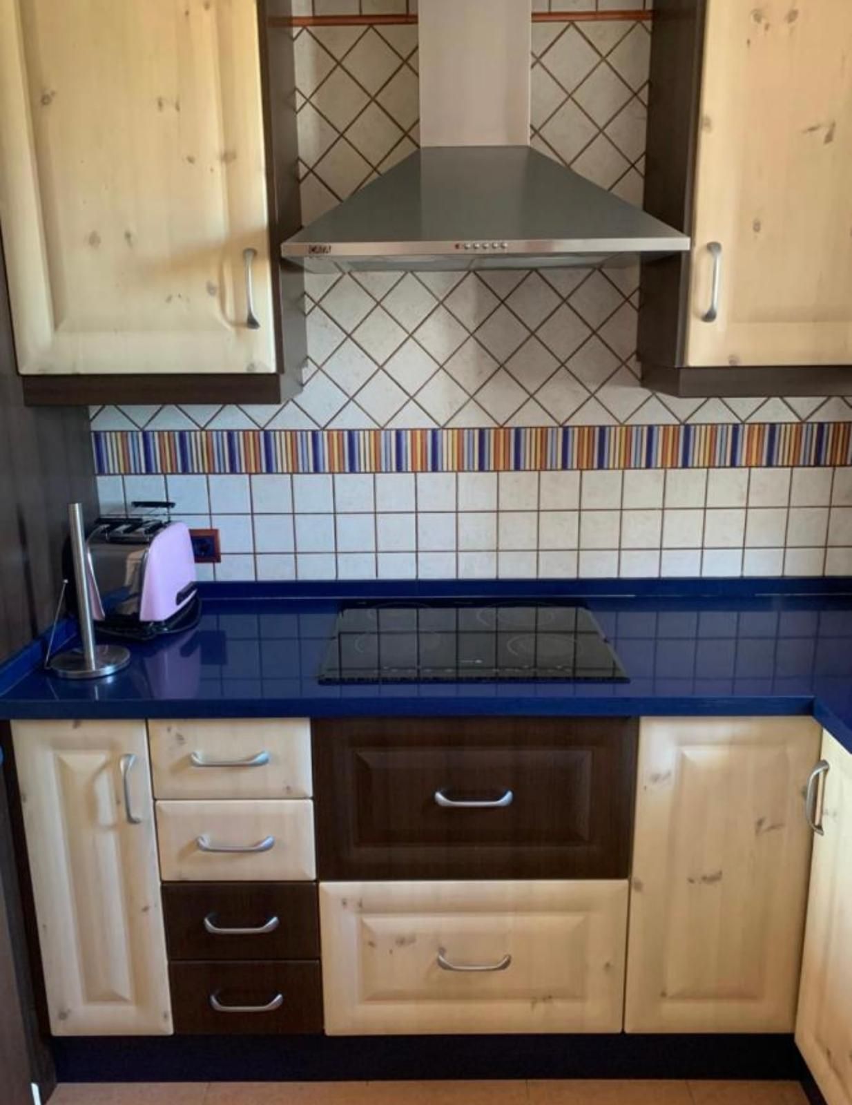 Cocina con encimera azul, gabinetes de madera clara y detalles en marrón oscuro; ventilación superior y protector contra salpicaduras de azulejos.
