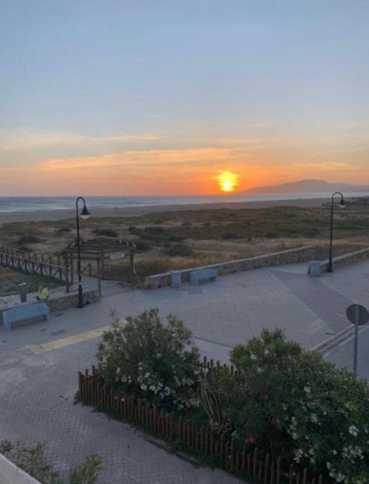 Puesta de sol sobre una playa con pasarelas de madera, farolas y arbustos en primer plano.