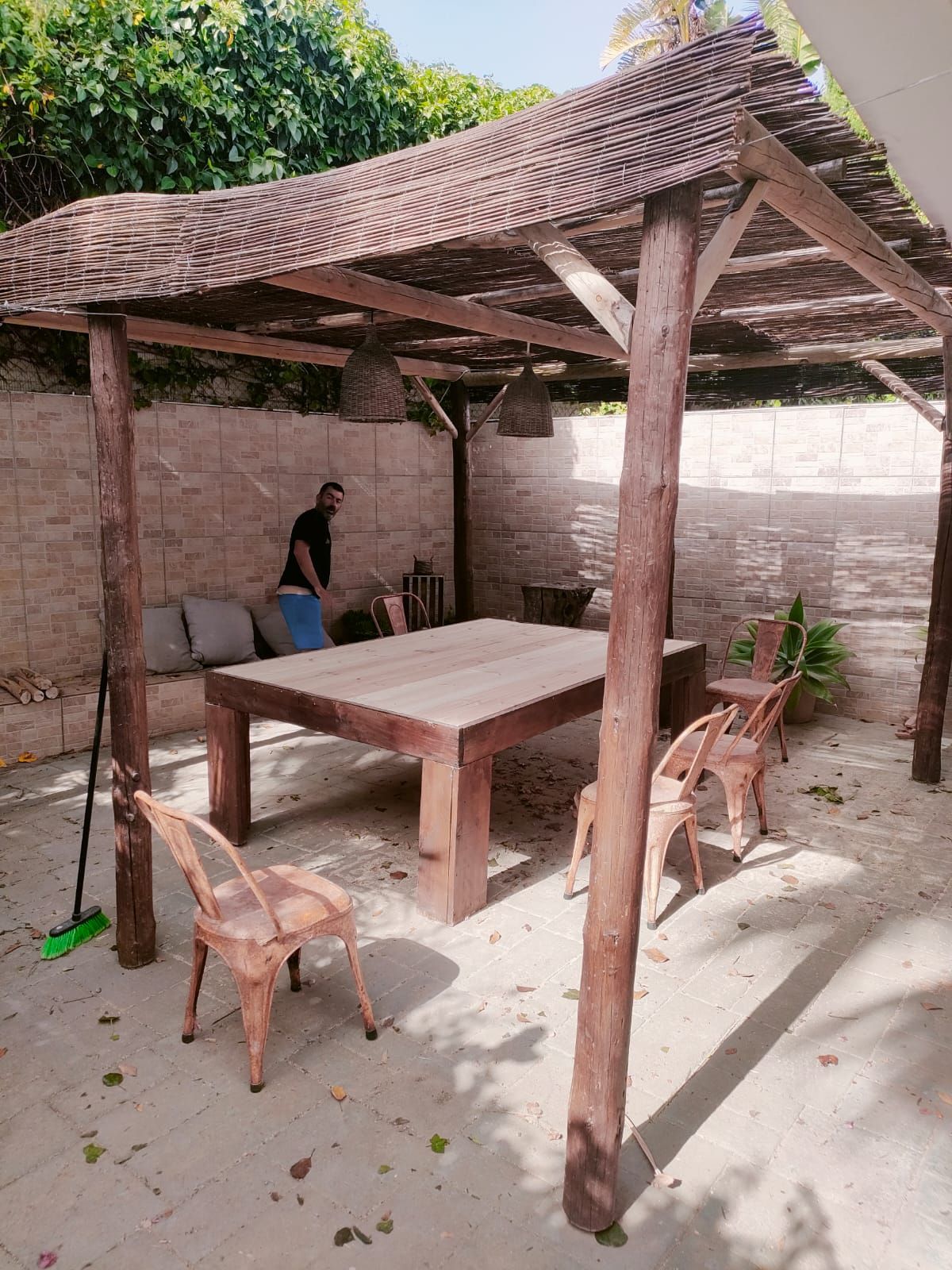 Comedor al aire libre con mesa de madera, sillas y pérgola. Hombre de pie junto a la mesa, tonos marrón claro.