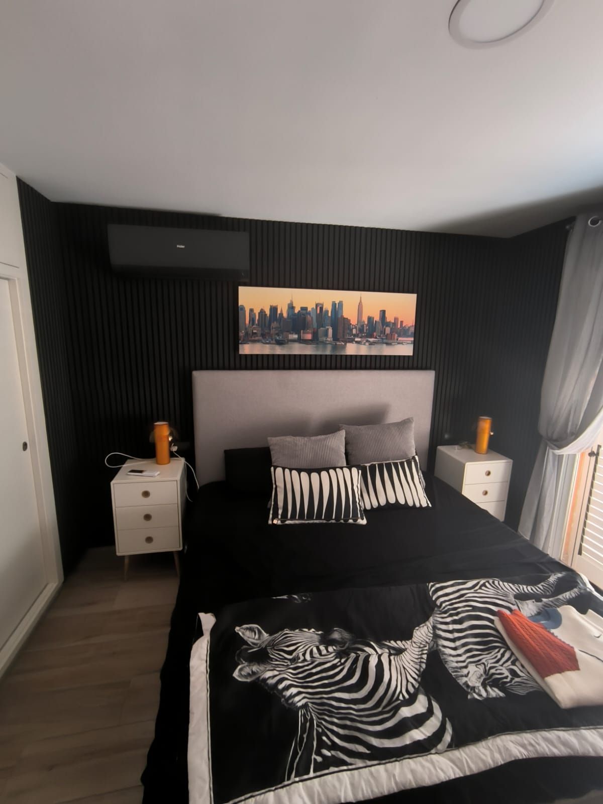 Dormitorio con pared con acento negro, cama, mesitas de noche y una pintura del horizonte de la ciudad.