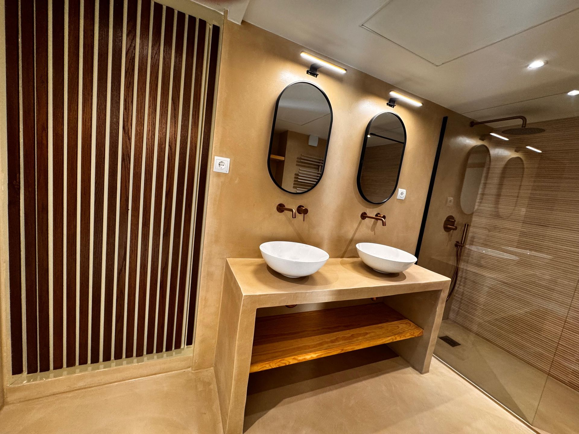 Baño con dos lavabos, espejos ovalados y una pared de listones de madera. Tonos tierra y diseño moderno.