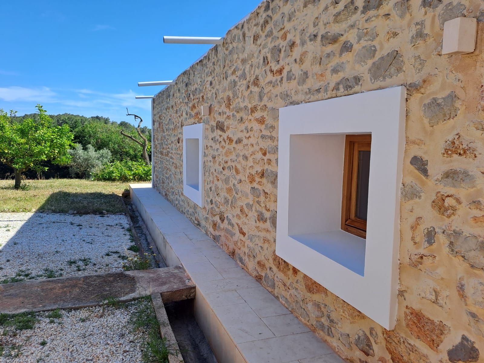 Exterior de edificio de piedra con ventanas de marco blanco, un banco de hormigón y un camino de grava, bajo un cielo azul.