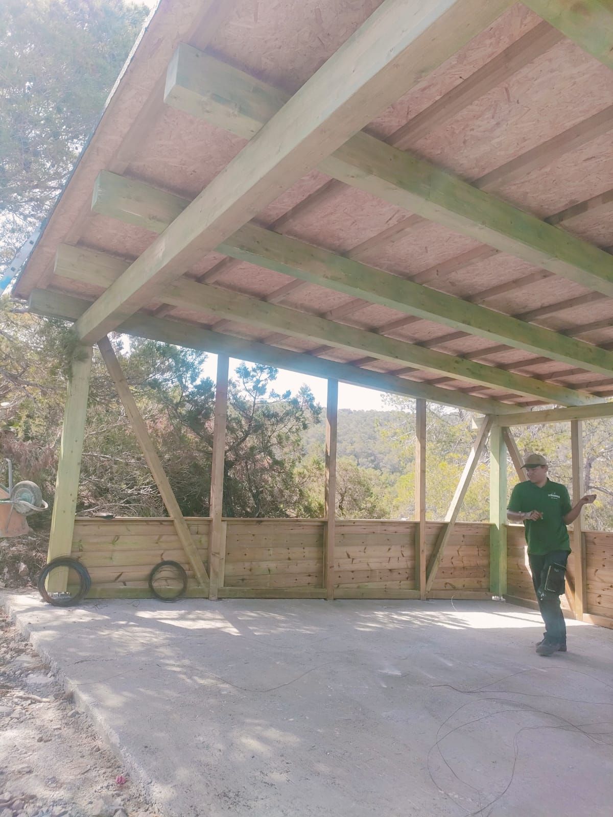 Cochera de madera, abierta por un lado. Un hombre vestido de verde está dentro. Suelo de hormigón, rodeado de naturaleza.