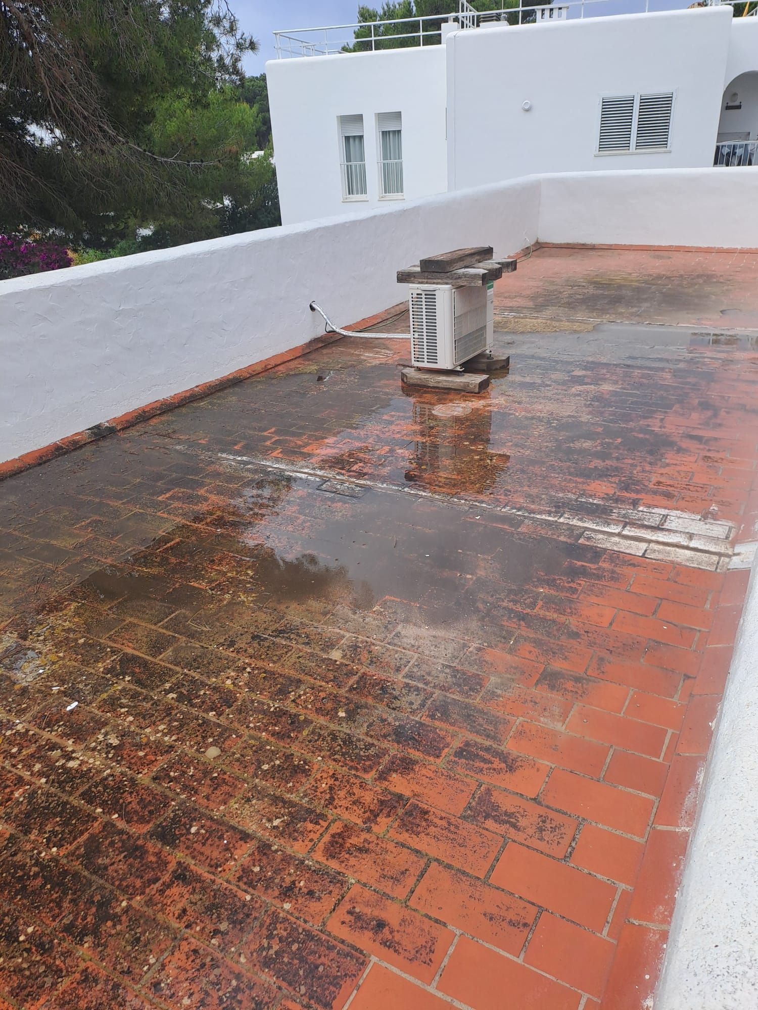 Tejado plano de tejas rojas con agua estancada y un aparato de aire acondicionado; edificio blanco al fondo.
