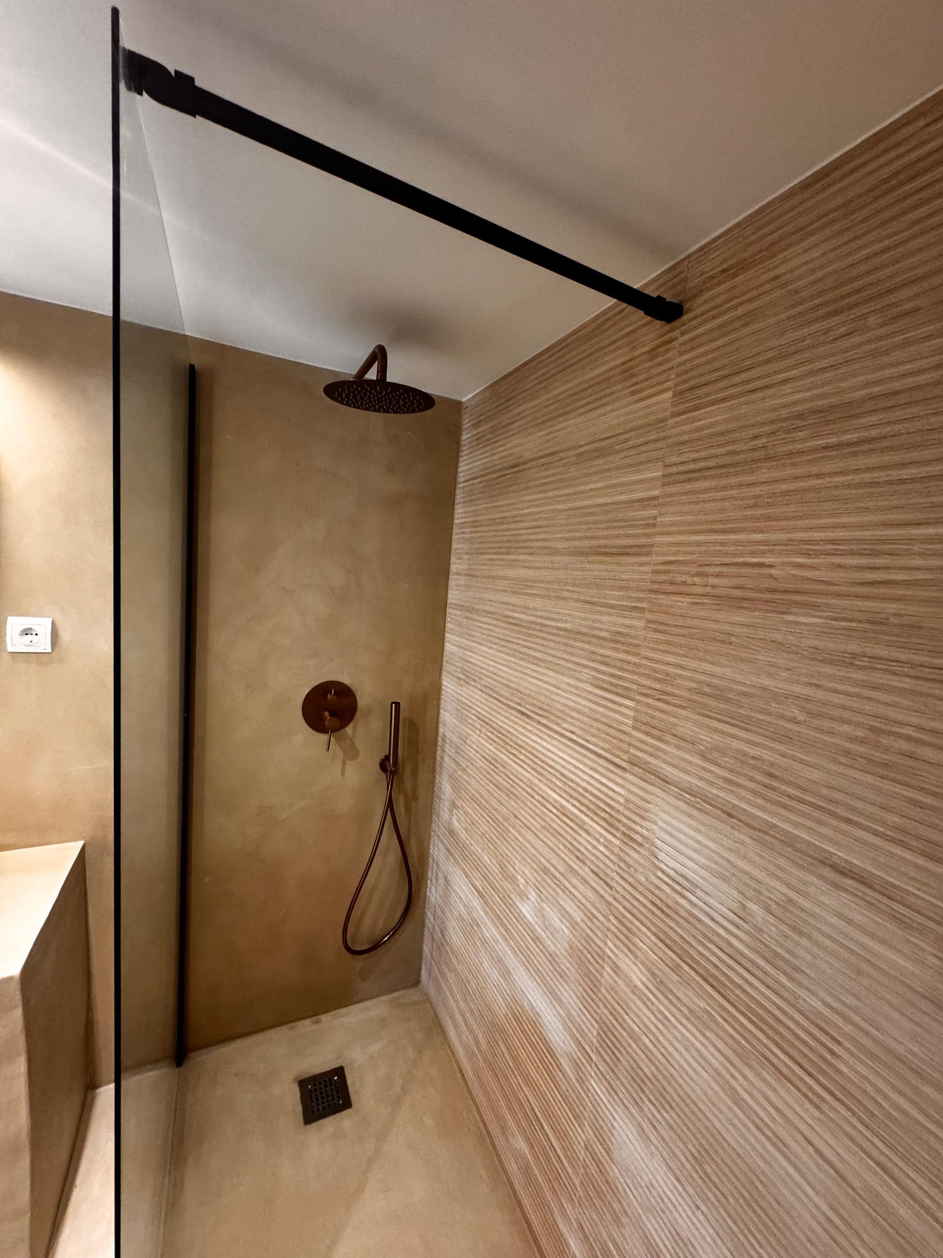 Ducha con paredes texturizadas de color marrón, accesorios oscuros y panel de vidrio.