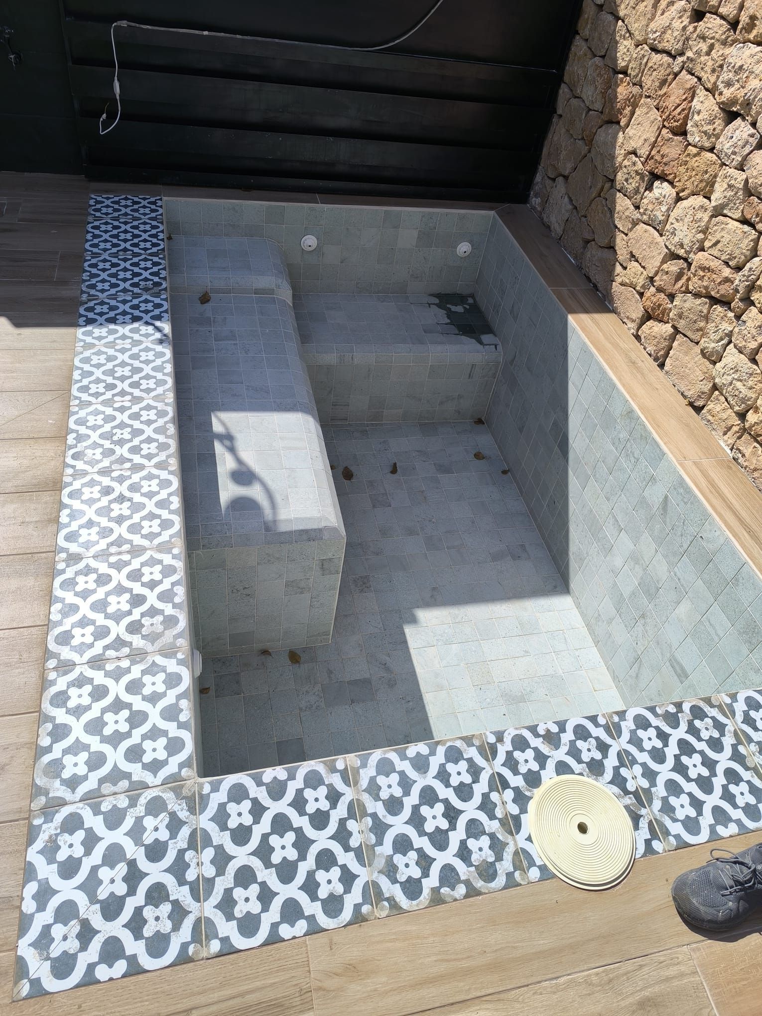 Piscina de inmersión vacía con azulejos, escalones, rodeada de azulejos con borde decorativo y una pared de piedra.