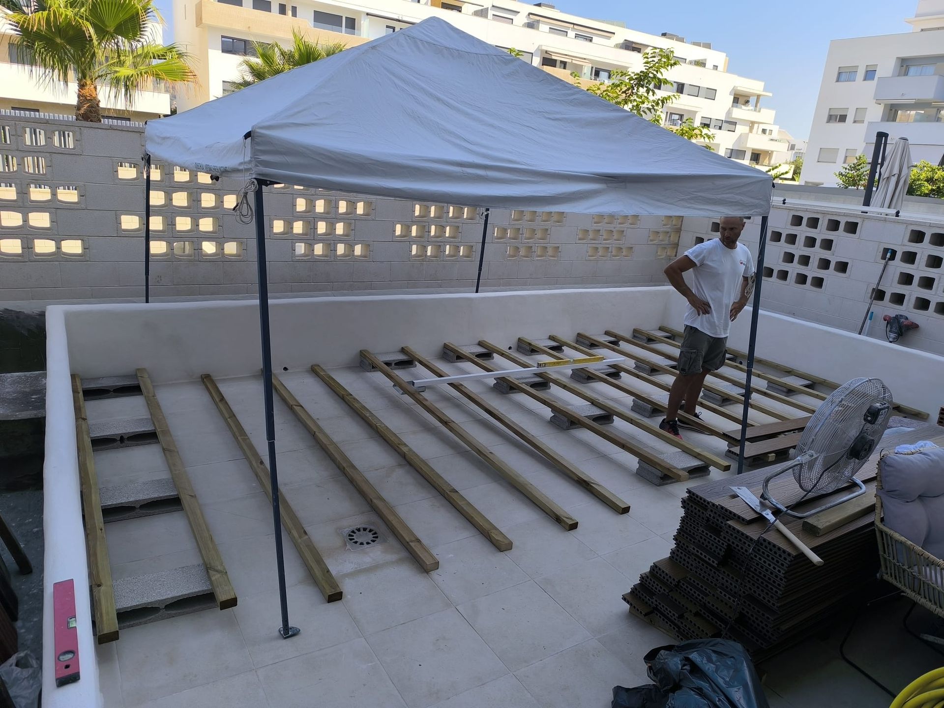 Hombre construyendo una terraza bajo un dosel blanco. Vigas de madera en un patio de hormigón, fondo de pared blanca.