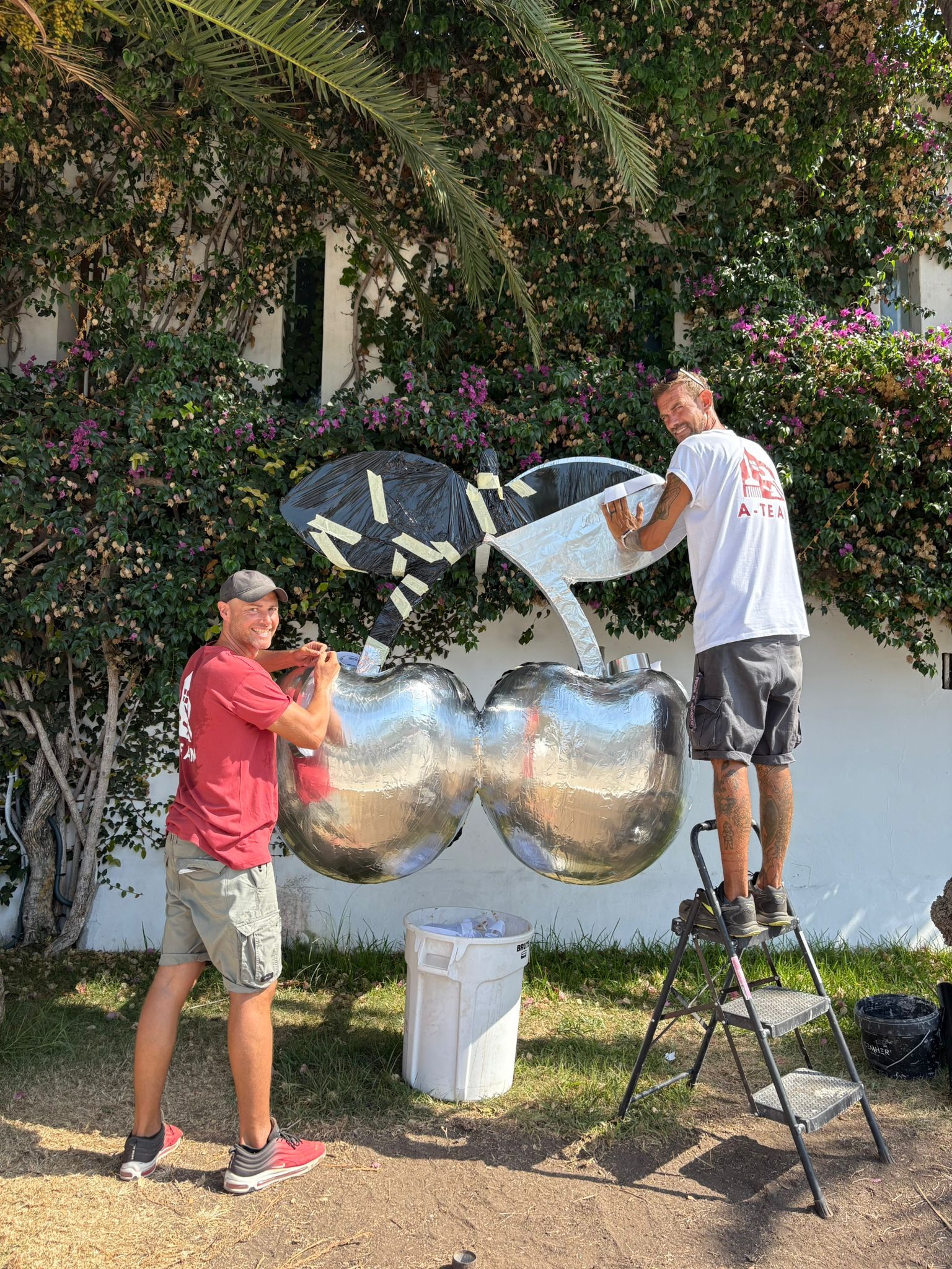Dos hombres instalan una gran decoración con globos de color cereza plateados al aire libre, cerca de un seto frondoso y una pared blanca.