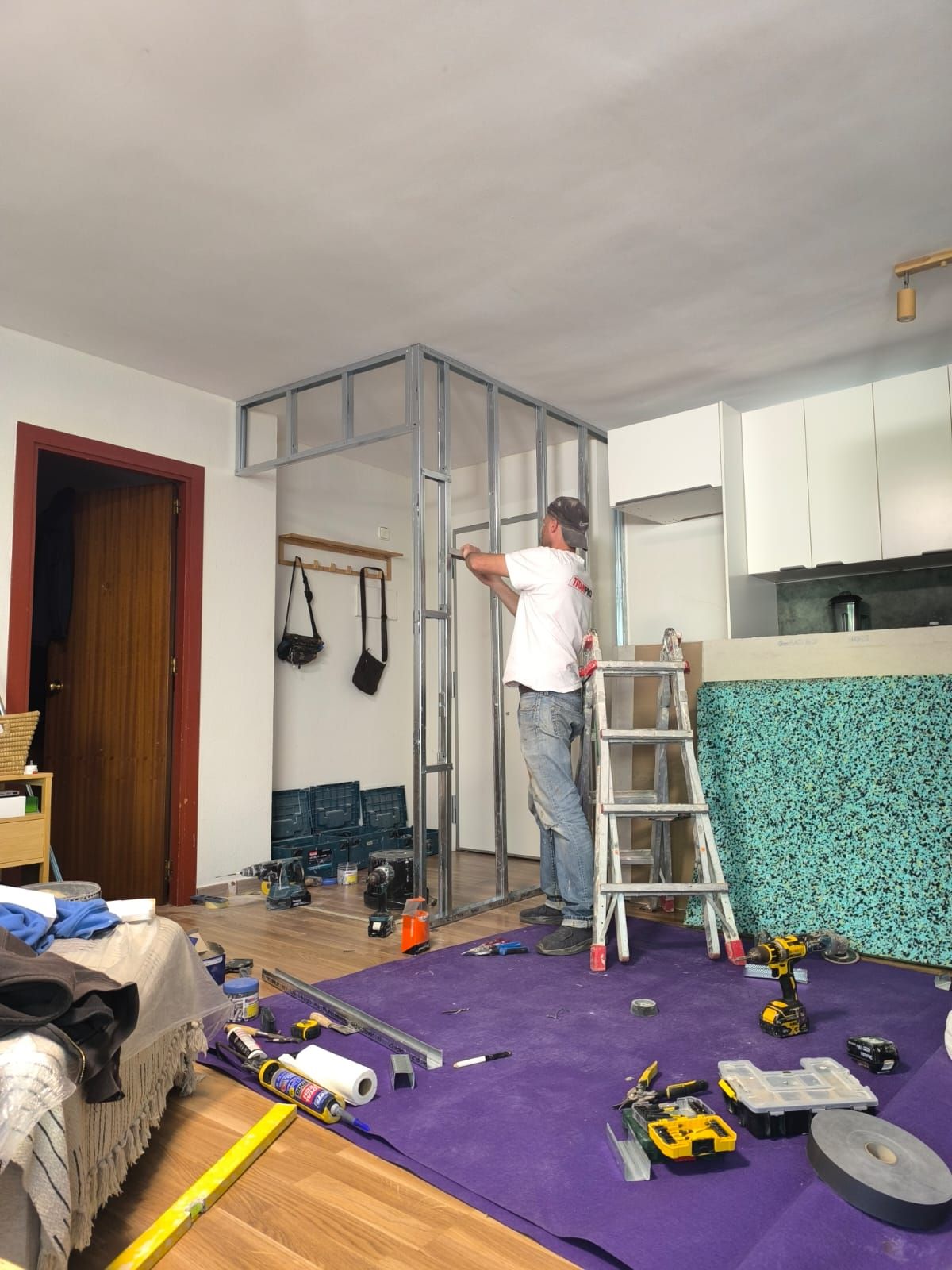 Hombre construyendo una pared de estructura metálica en un interior. Herramientas y escalera sobre suelo morado.