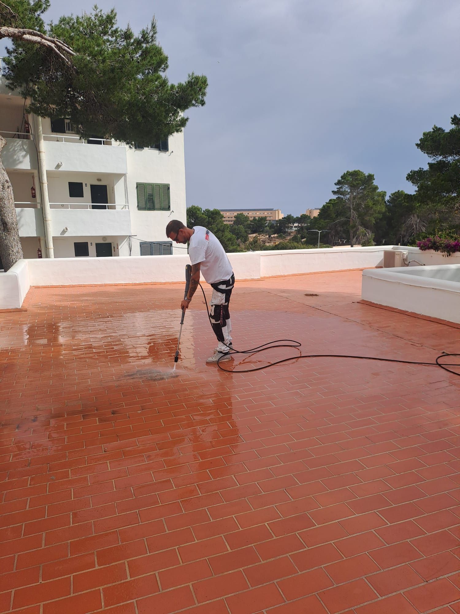 Un hombre lava a presión un techo de tejas rojas con un edificio al fondo bajo un cielo parcialmente nublado.