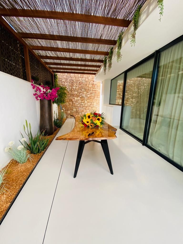 Comedor al aire libre con mesa de madera, girasoles y plantas bajo techo de pérgola.