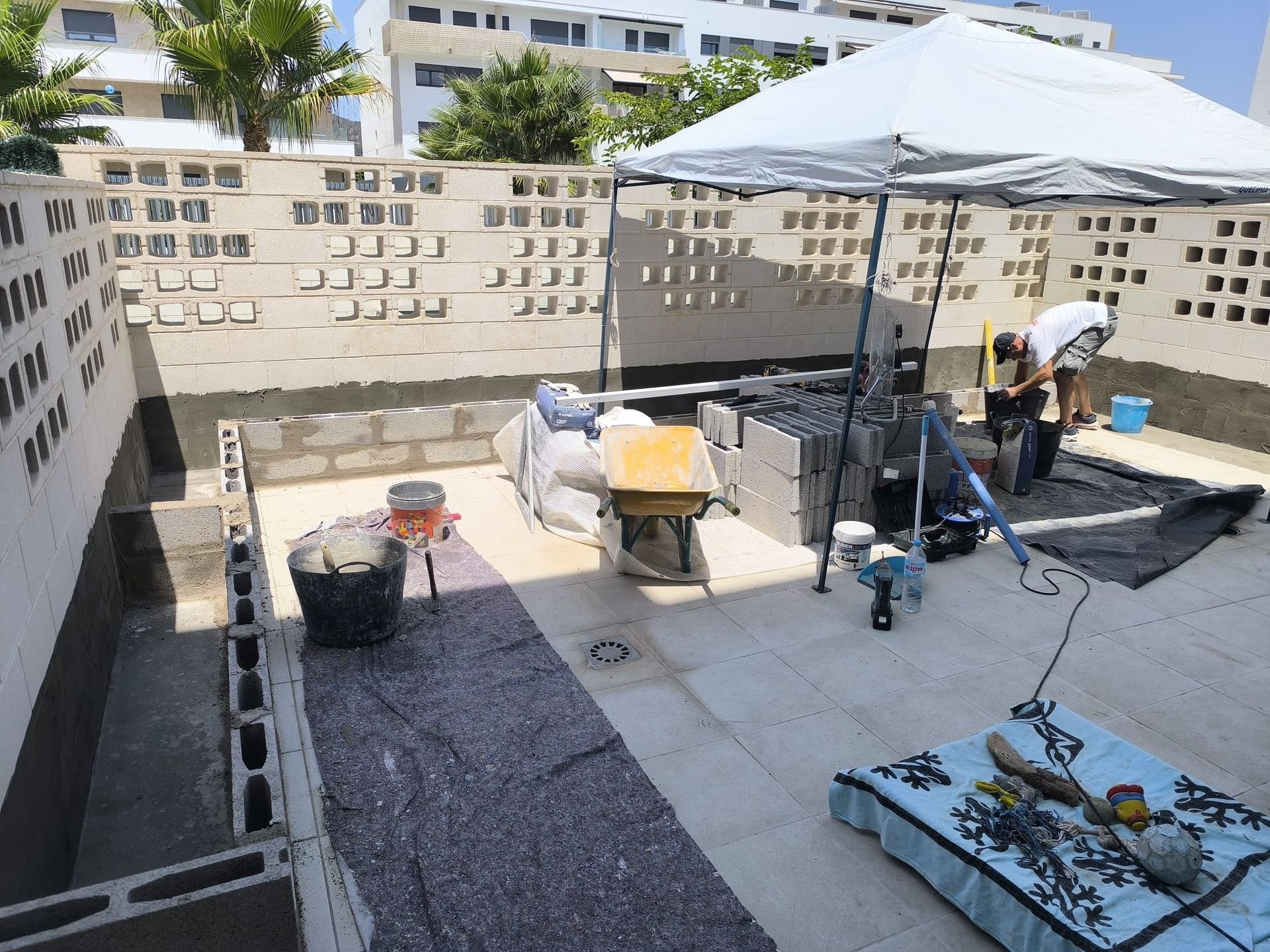 Escena de construcción: Trabajadores construyen un muro con bloques, mortero y herramientas en un tejado debajo de una tienda de campaña.