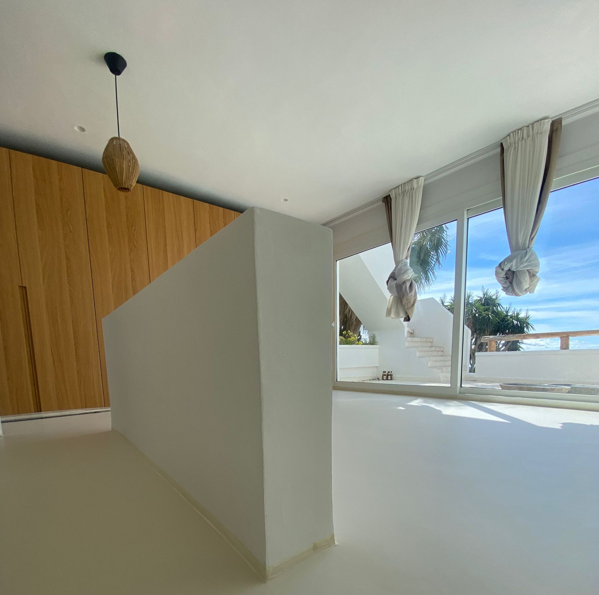 Habitación luminosa con paredes y suelo blancos. Revestimiento de madera, ventanal con vistas y lámpara colgante.