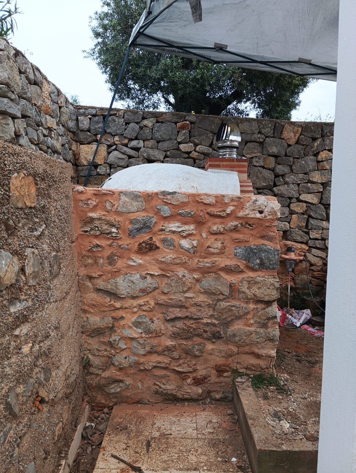 Muros de piedra encierran una zona exterior con un horno de piedra y un árbol al fondo.