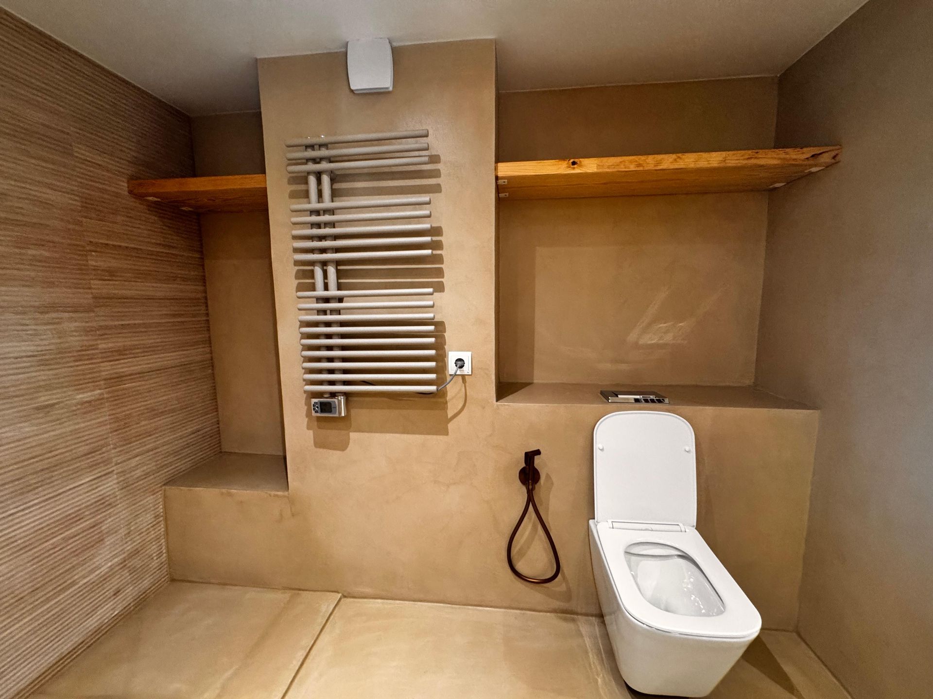 Baño con inodoro blanco, toallero calefaccionado, estante de madera y paredes de color tostado.